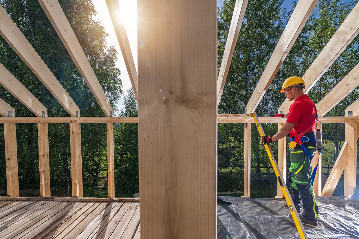 A worker on a ladder using a level to check the alignment of a wooden roof frame.