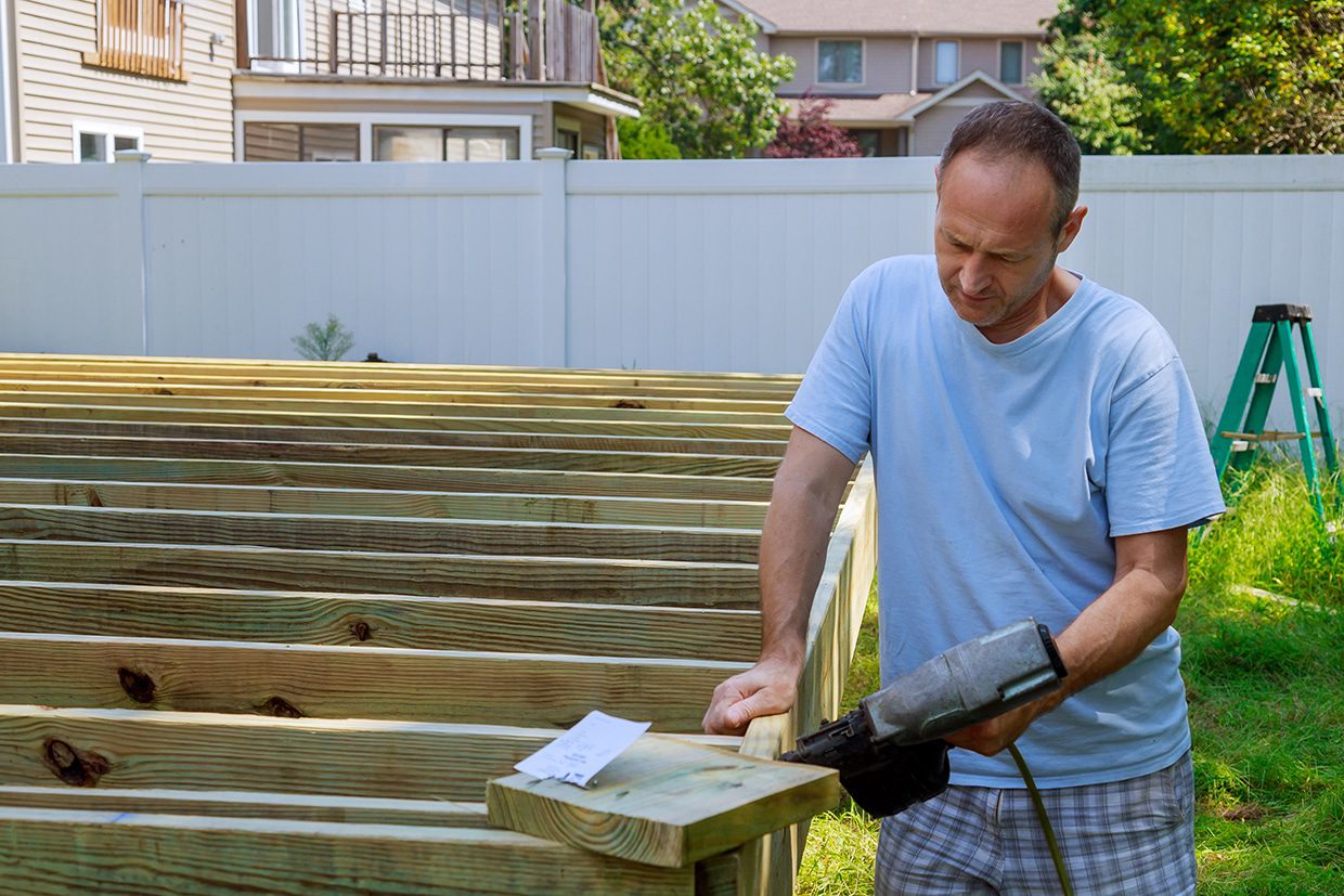 Builder using a pneumatic nail gun to secure pressure-treated joists for a new deck structure.