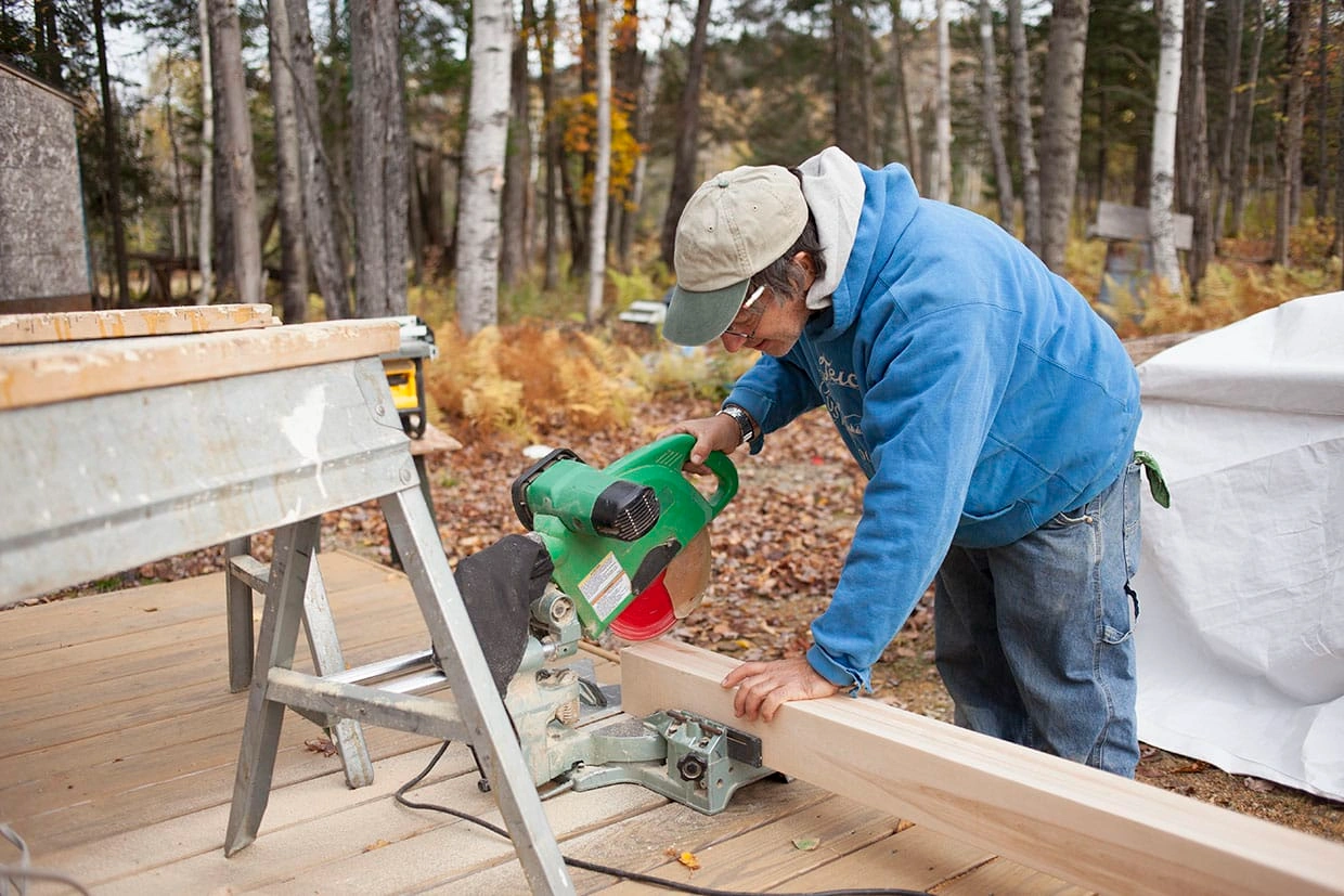 A carpenter using a green miter saw to cut a thick wooden beam for a custom deck project.