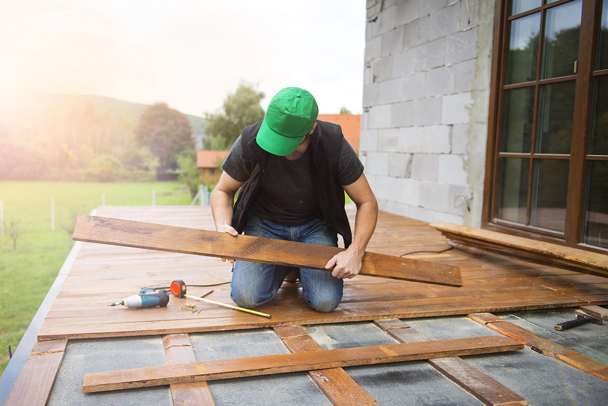 A man kneeling on a wooden deck inspecting a loose plank for potential replacement.