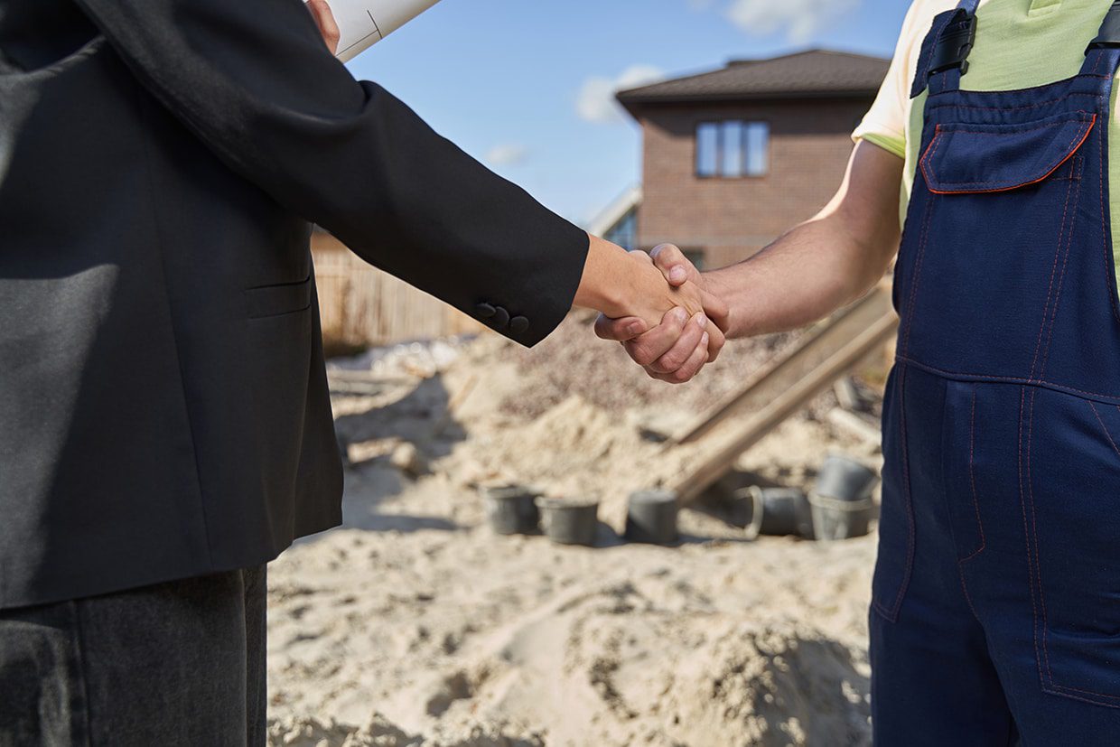 Close-up of a homeowner in a suit shaking hands with a contractor in blue overalls at an active construction site.