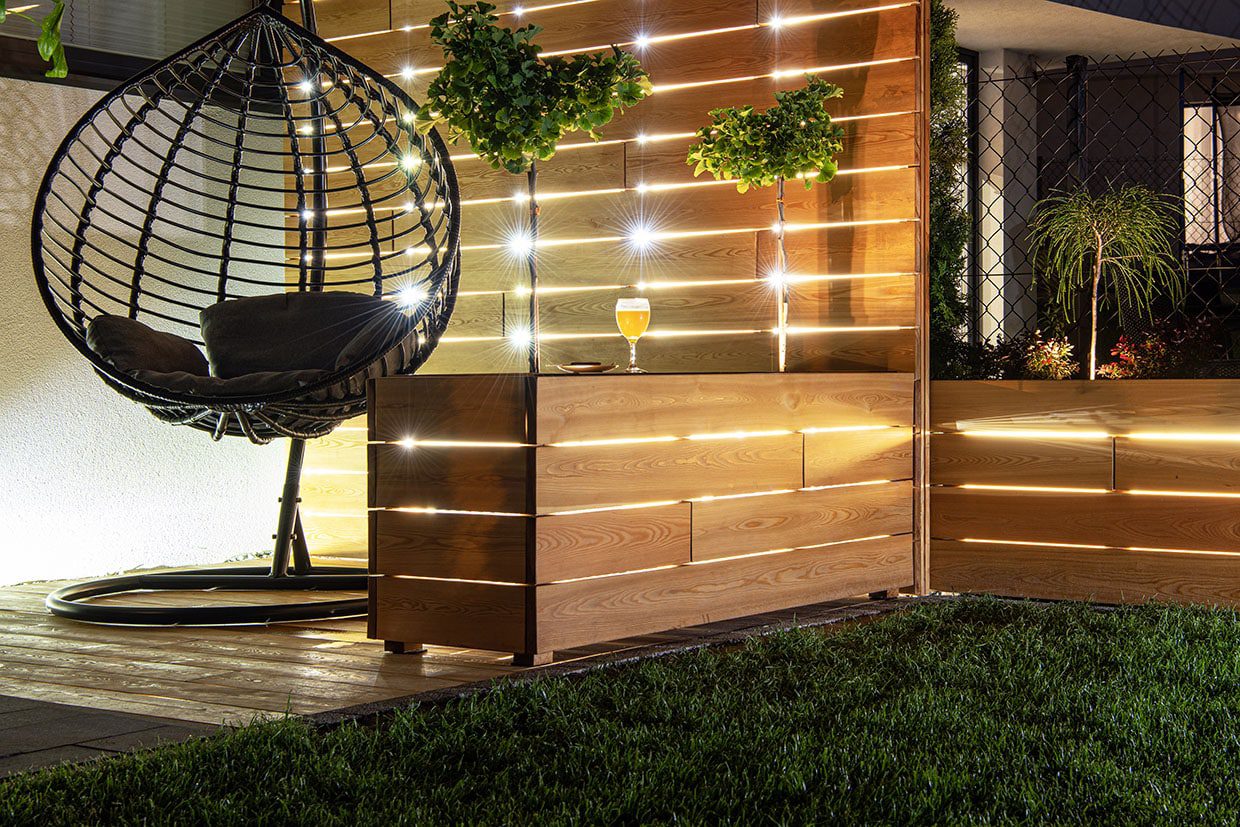 Illuminated wooden privacy wall with a hanging egg chair, showcasing creative deck resurfacing ideas.