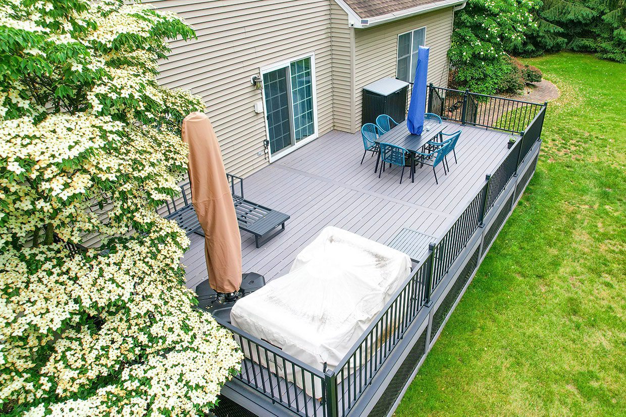 High-angle view of a grey composite deck with dining furniture, bordered by a large flowering dogwood tree and beige house siding.