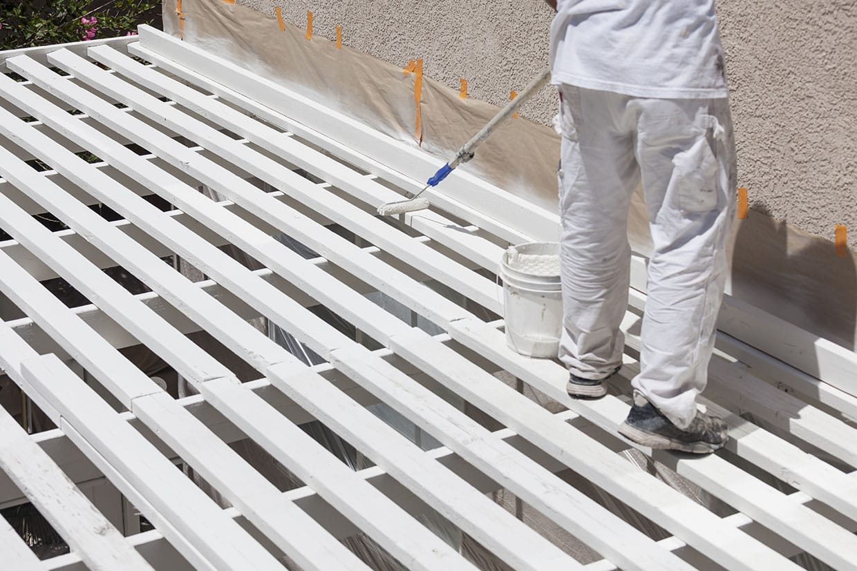 Painter applying white paint to the top beams of a wooden pergola structure.
