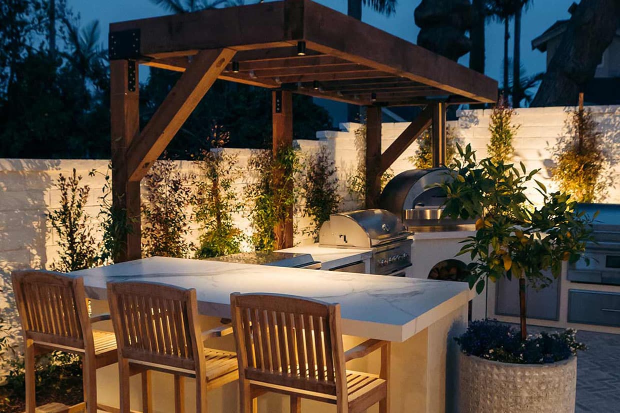 Illuminated outdoor bar and pizza oven area at night under a wooden pergola structure.