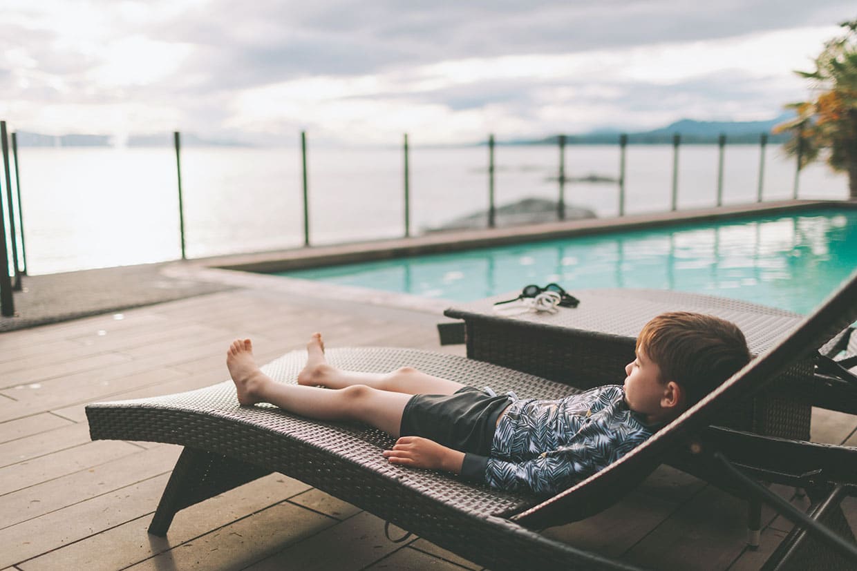 Child relaxing on a lounge chair on a poolside deck with glass railings overlooking the ocean.