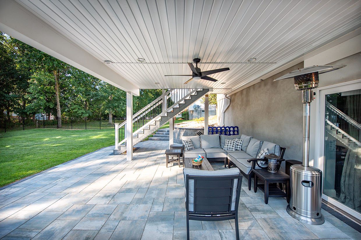 A paved patio living area situated beneath a white second-story deck ceiling with a fan and stairs leading up.