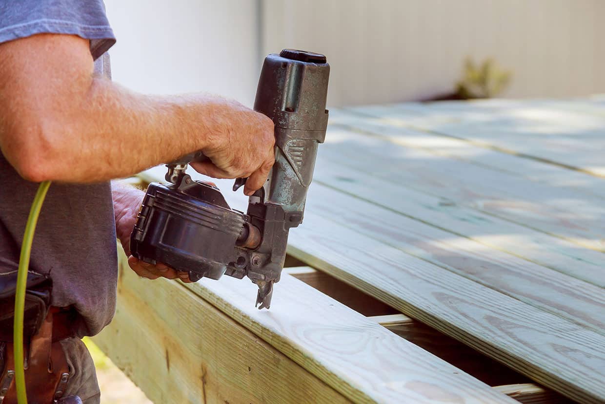 Worker using a pneumatic nail gun to fasten new pressure-treated lumber during a deck replacement.