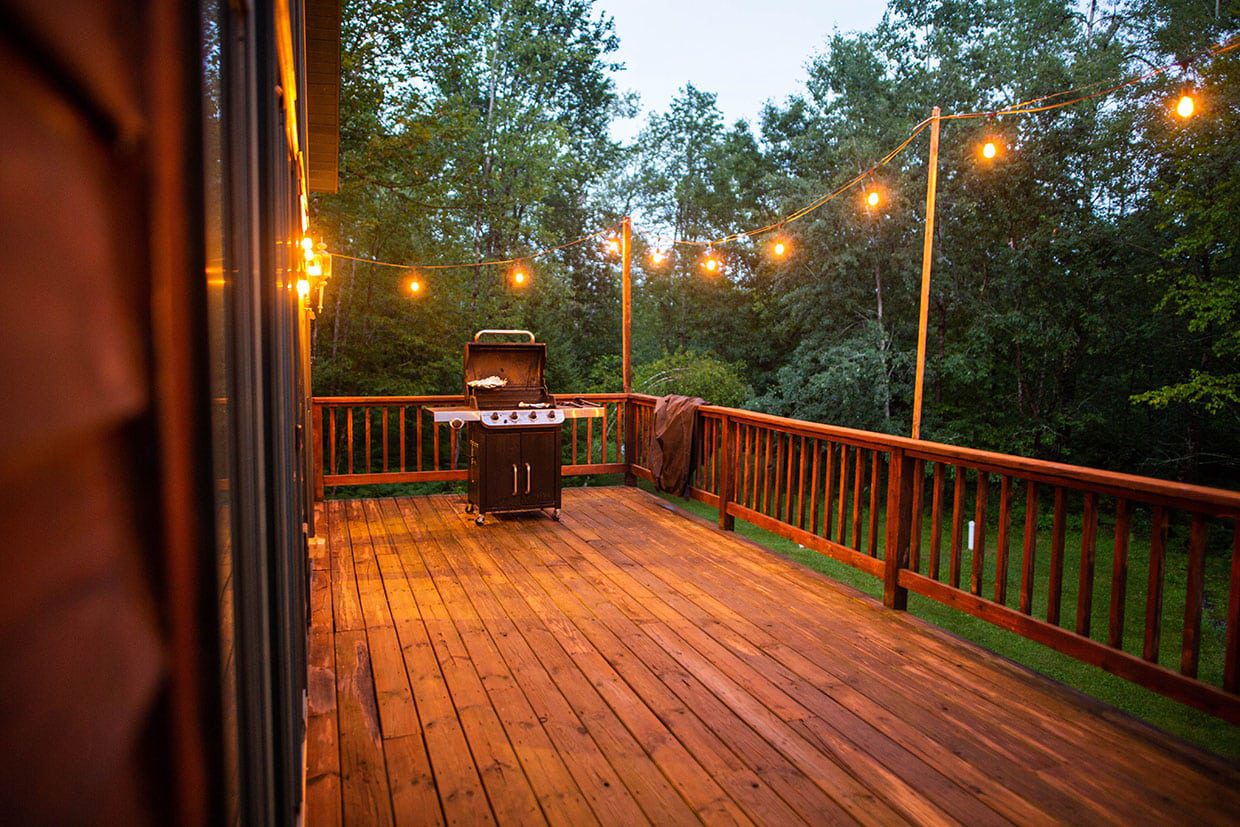 A spacious wooden deck illuminated by warm string lights at twilight with a covered grill, illustrating how to add value to your home with a new deck.