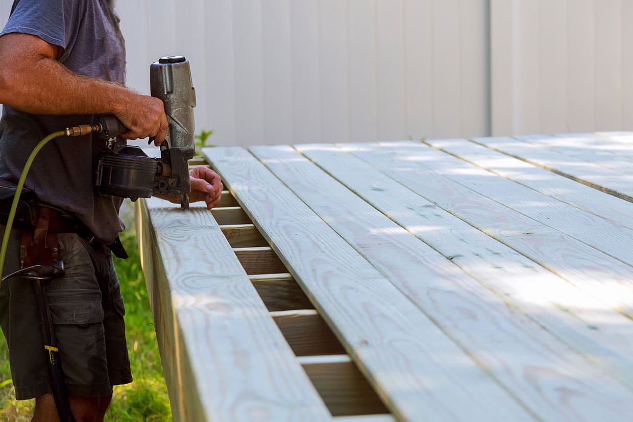 A contractor secures light-colored pine boards with a pneumatic nailer, illustrating the installation process for real wood decks.