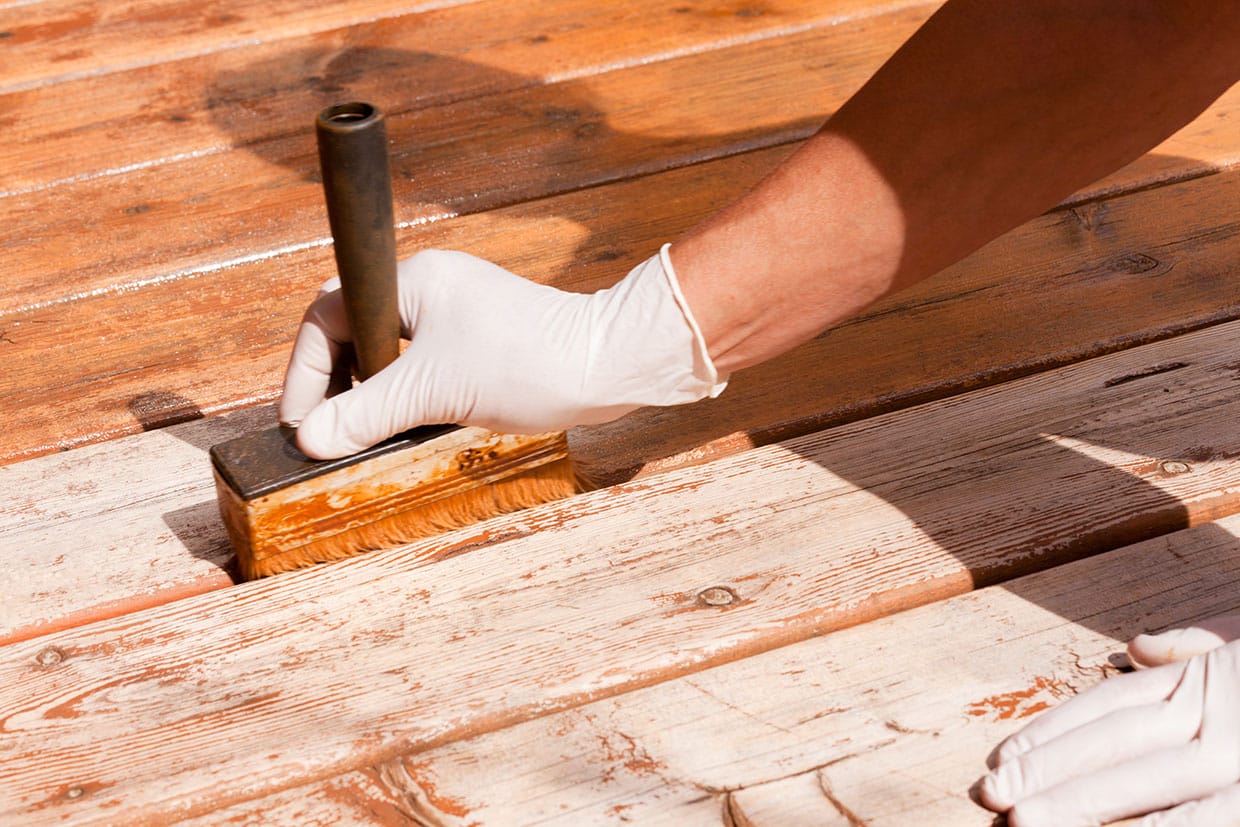 Close-up of a hand applying reddish-brown stain to weathered wood during a deck resurfacing project.
