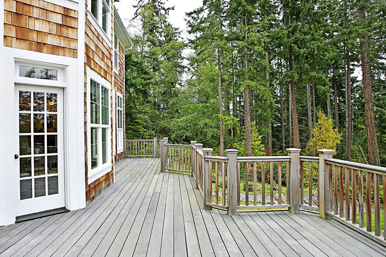 A large, weathered gray deck attached to a home with cedar siding, showing the natural aging of wood without regular sealing.