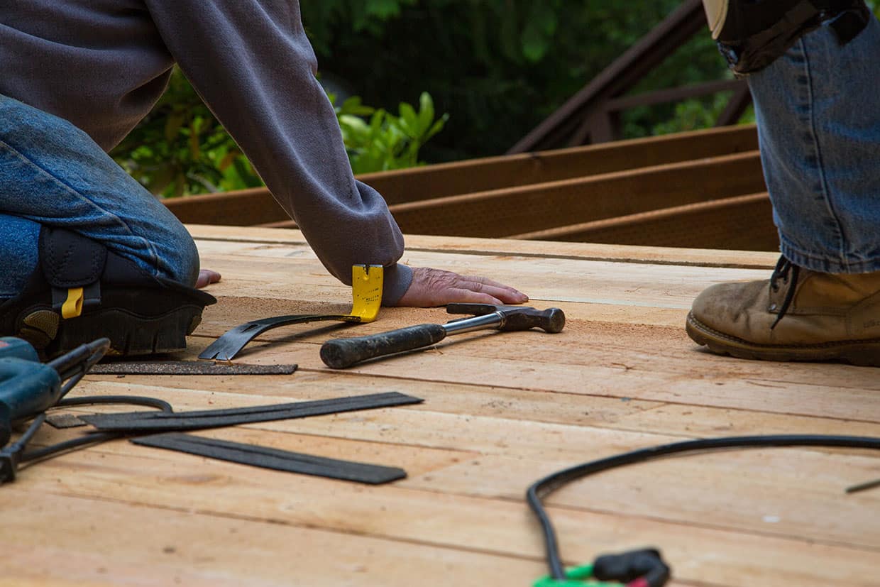 Two workers kneeling on unfinished wooden planks with hammers and pry bars, performing manual wood deck maintenance or repairs.