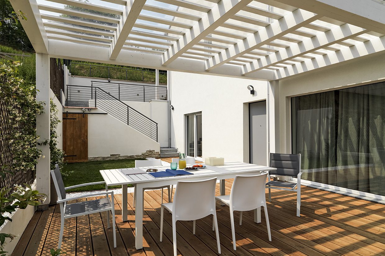 White dining set on a wooden deck located in a courtyard with a white pergola structure overhead.