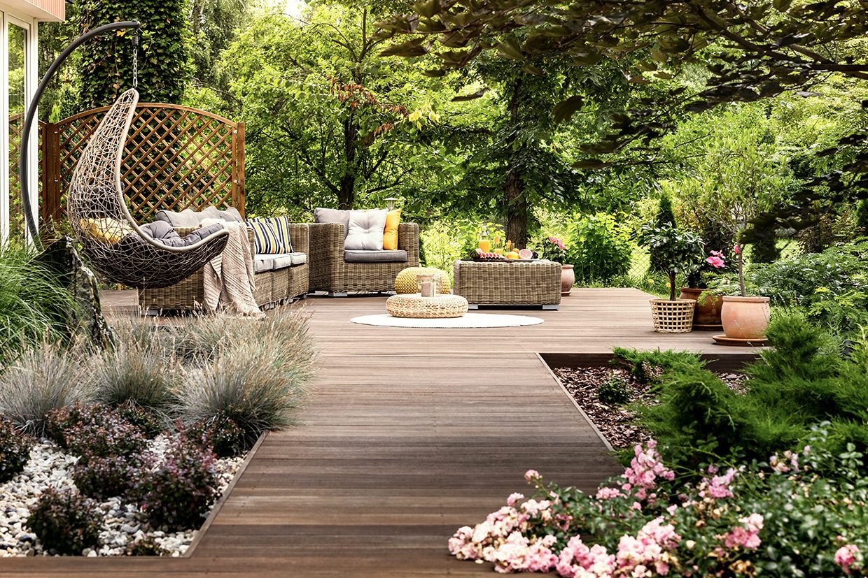 A spacious wooden deck walkway leading to a wicker seating area and hanging chair surrounded by lush green trees.