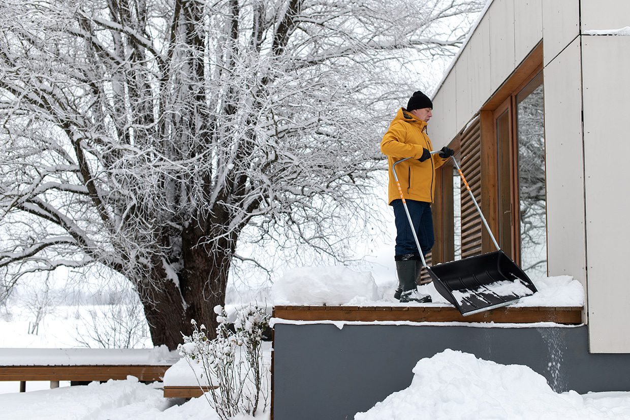 A person in a yellow jacket using a shovel to clear heavy snow, helping prepare deck for winter.