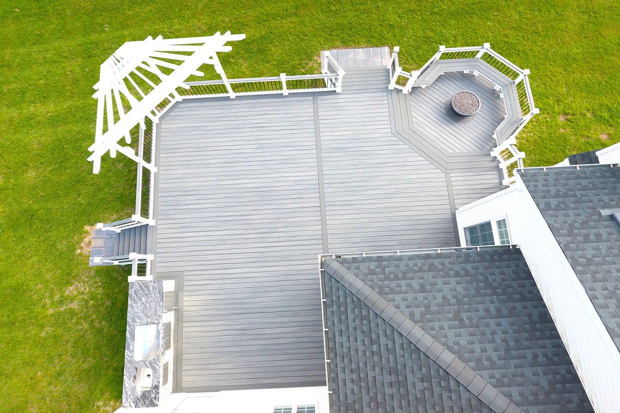 Side-by-side comparison of a covered white pergola deck versus an open composite deck, both overlooking a green lawn.