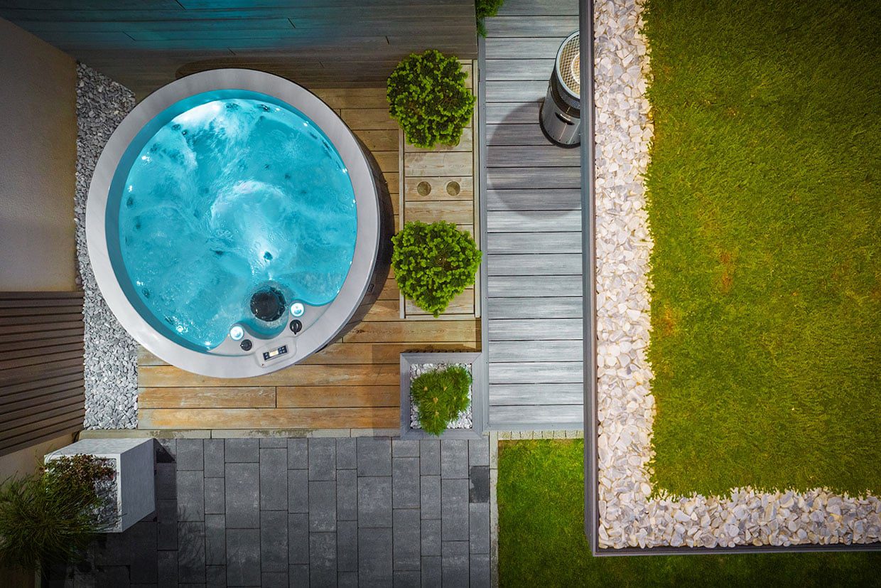 Top-down view of a round hot tub built into a wood and stone deck, accented with river rock borders and planter boxes for garden deck integration.