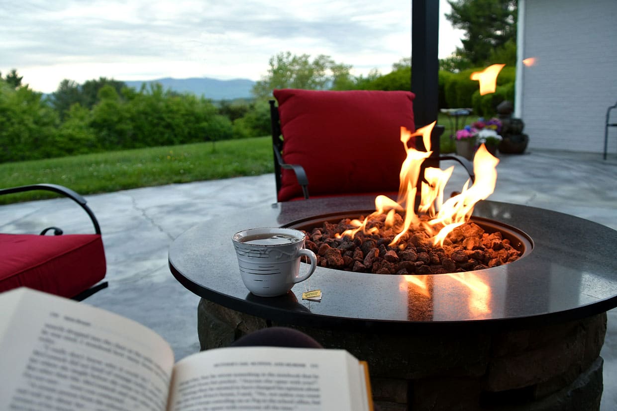 Close-up of a round gas fire pit with lava rocks on a concrete patio, featuring a tea mug and open book in the foreground to illustrate relaxing smart deck features.