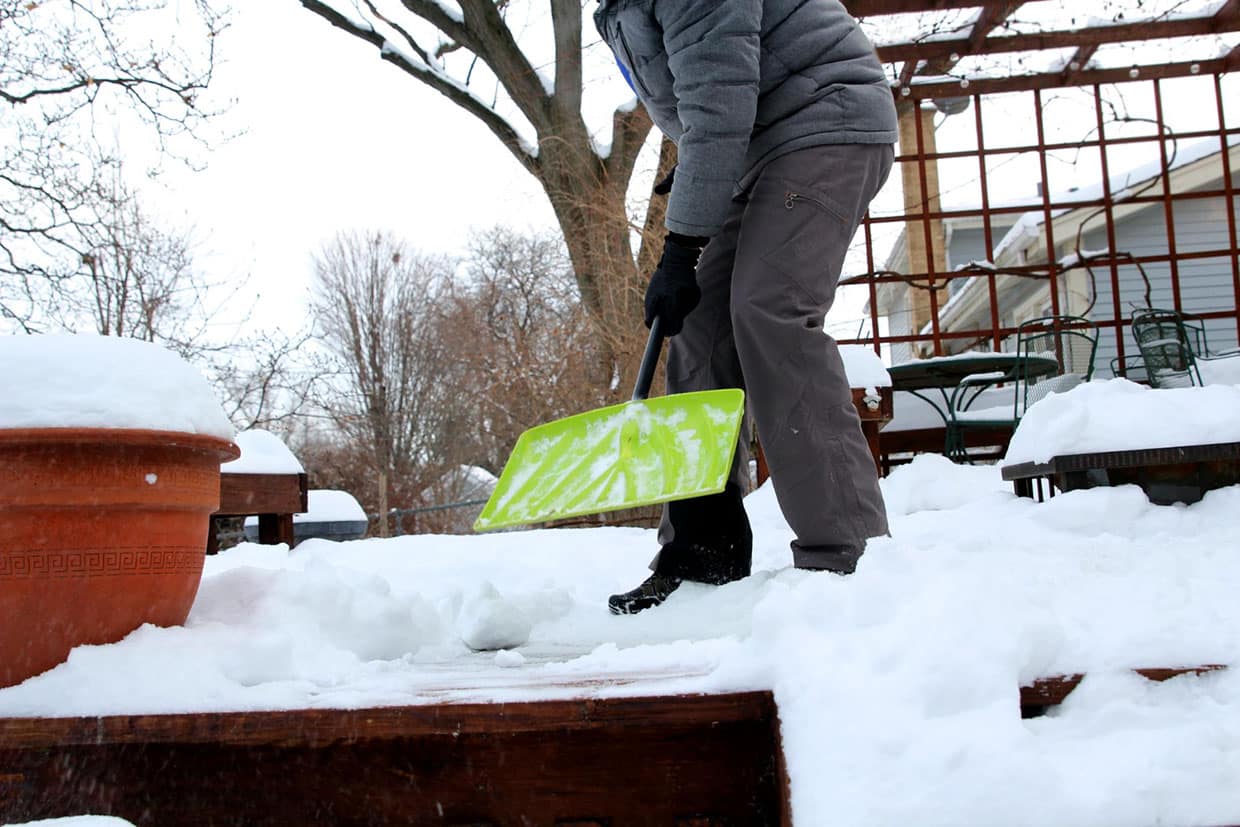 Person using a green shovel to clear snow from a wooden surface as part of year-round deck maintenance.