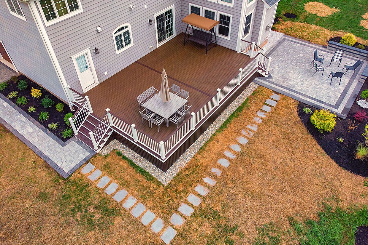 Aerial view of a home featuring a seamless deck and patio combination connected by a stone walkway and garden beds.
