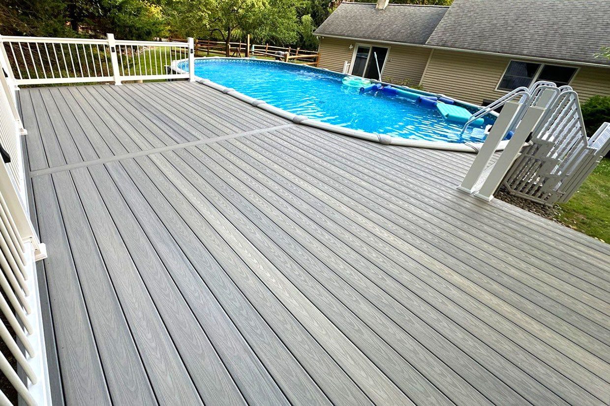 Large gray composite deck with white railings surrounding an above-ground swimming pool in a backyard.
