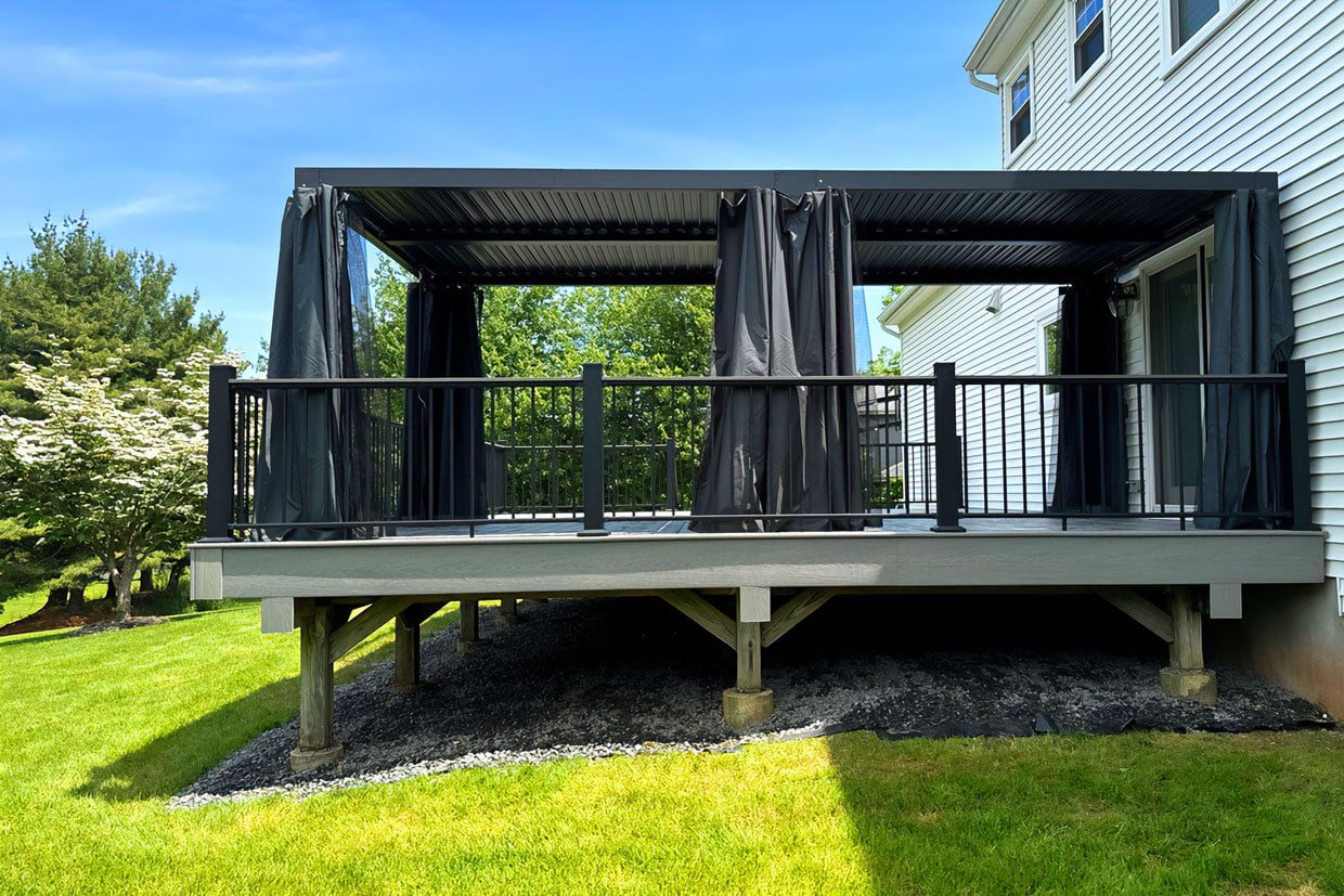 A raised grey composite platform featuring a black louvered pergola with privacy curtains, showcasing the best deck design for shade and style.