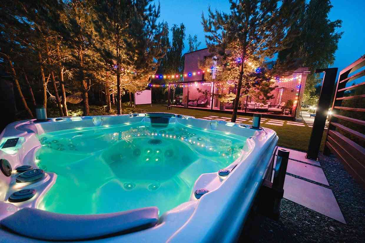 Illuminated large hot tub at night in a backyard setting with string lights and a house deck in the background.