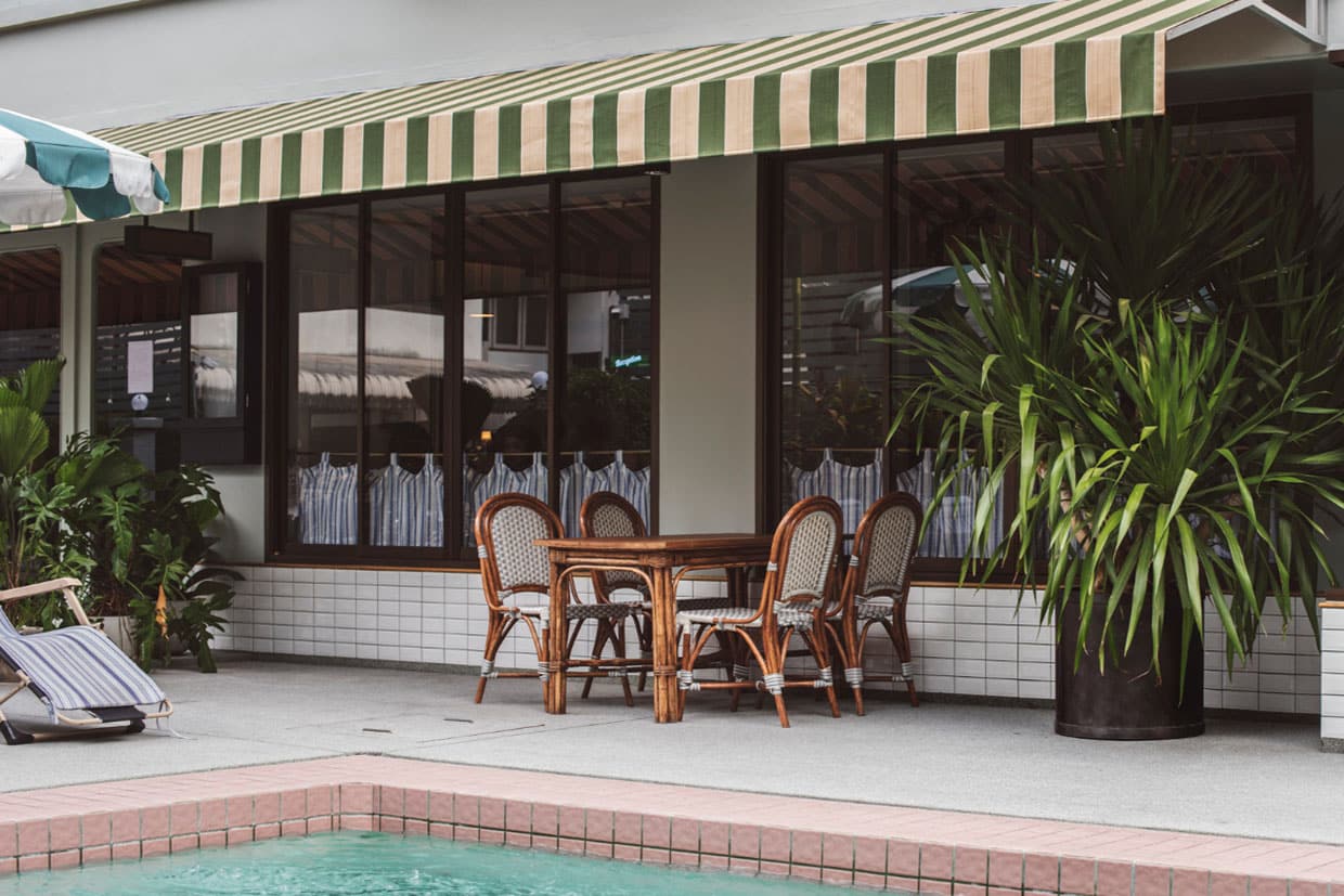 A green and beige striped fabric awning extends over a wooden dining table and rattan bistro chairs on a tiled patio next to a swimming pool, illustrating retro-style outdoor awning ideas.