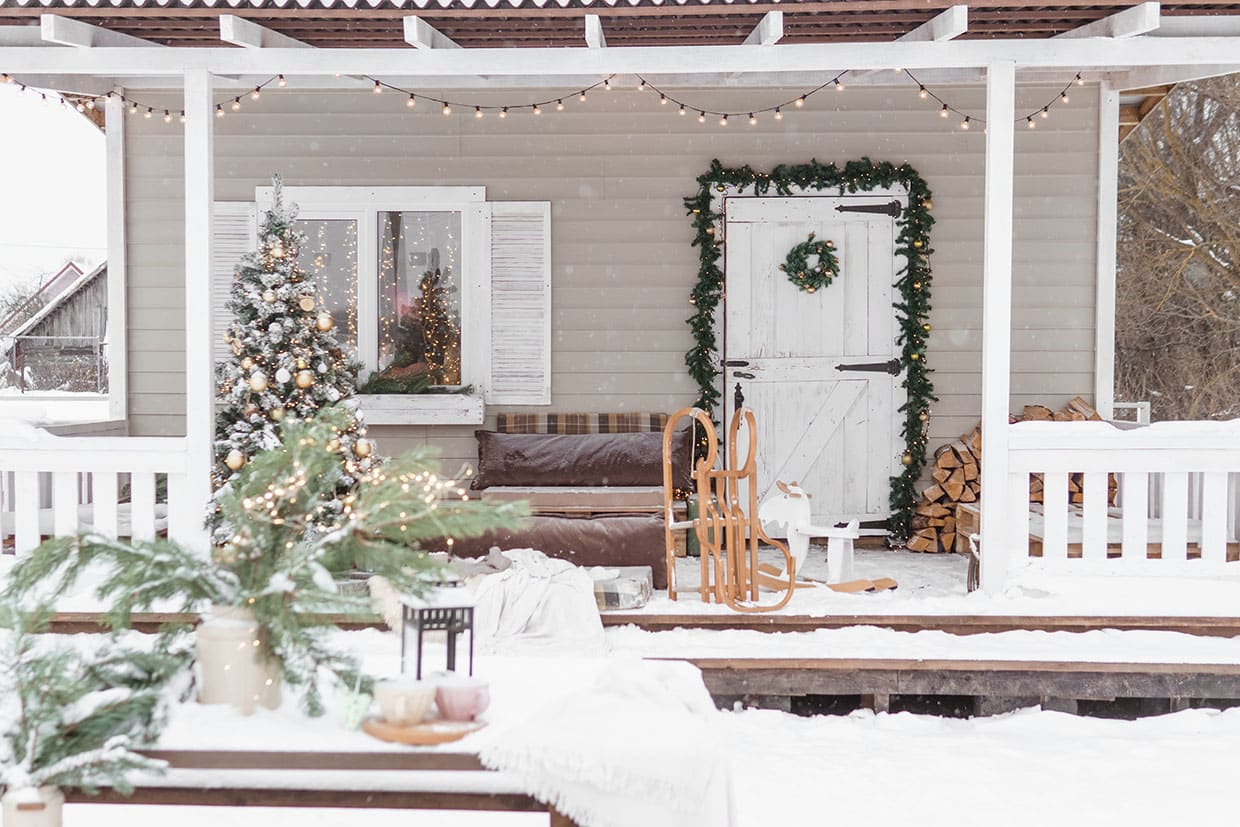 Snow-covered porch decorated with a Christmas tree, garland, wooden sled, and string lights as part of winter deck decorations.