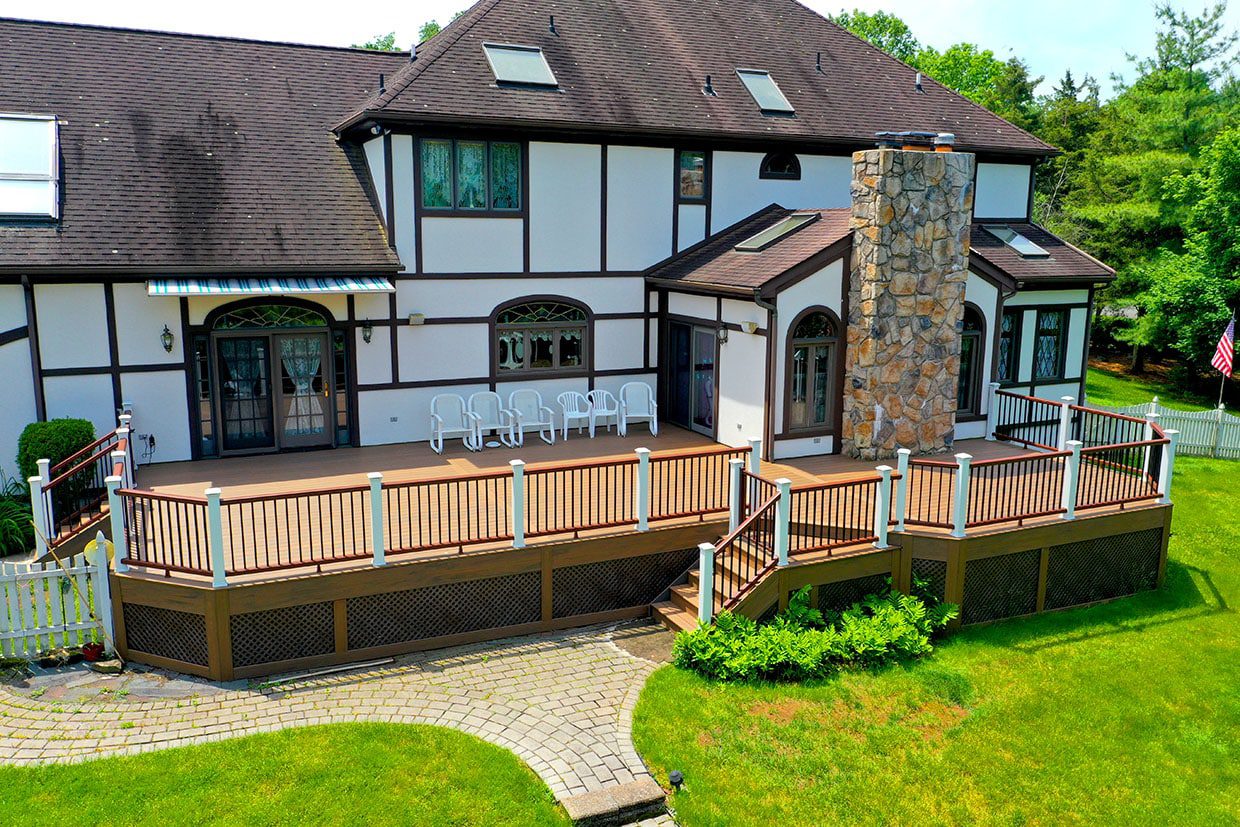 Expansive multi-level composite deck with white railings attached to a Tudor-style house with a stone chimney.