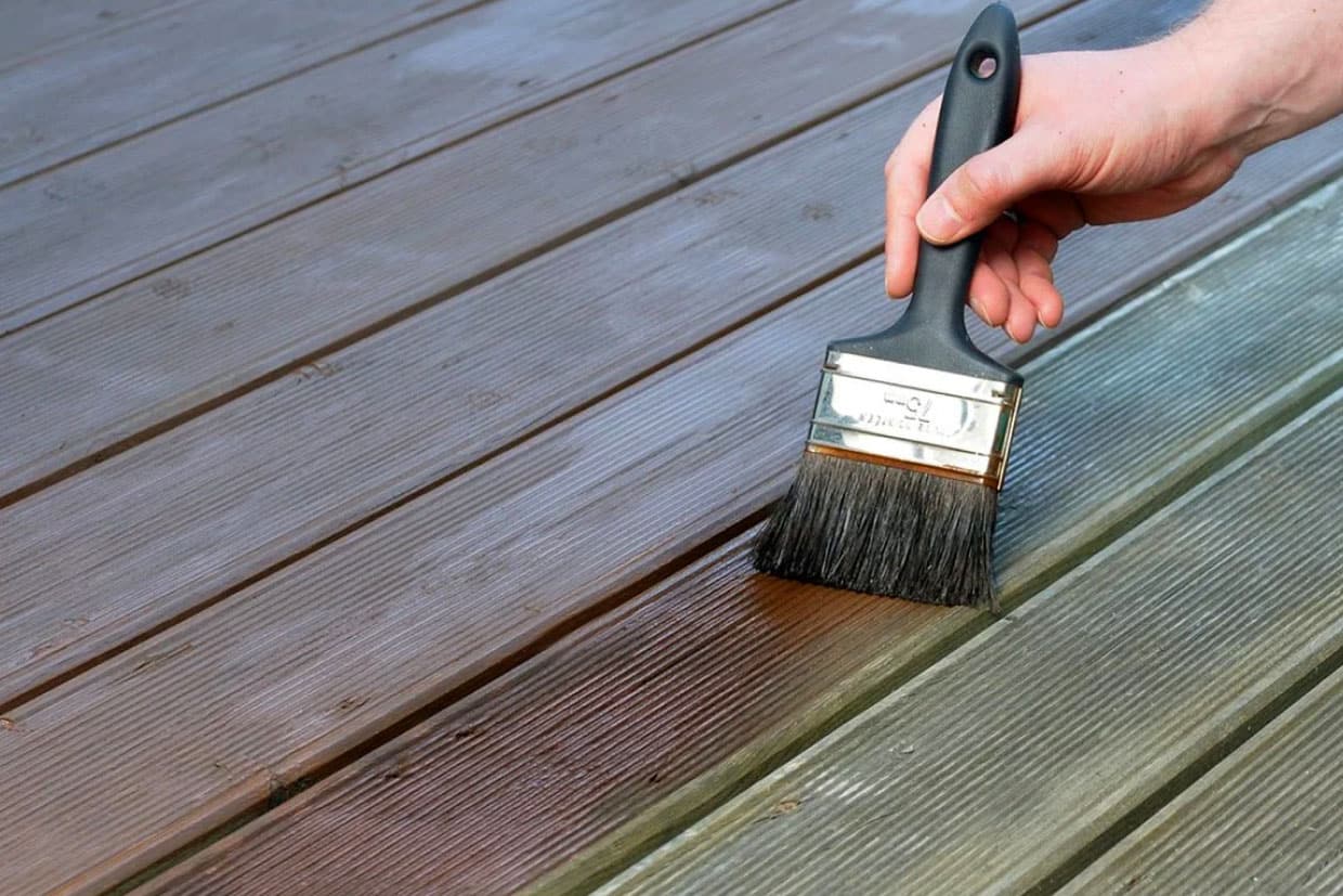 A hand holding a brush applying a liquid treatment to the wood-grain surface of the deck.