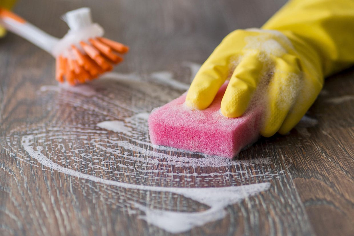 Close-up of a gloved hand using a sponge and eco-friendly deck cleaner to scrub a spot on the flooring.