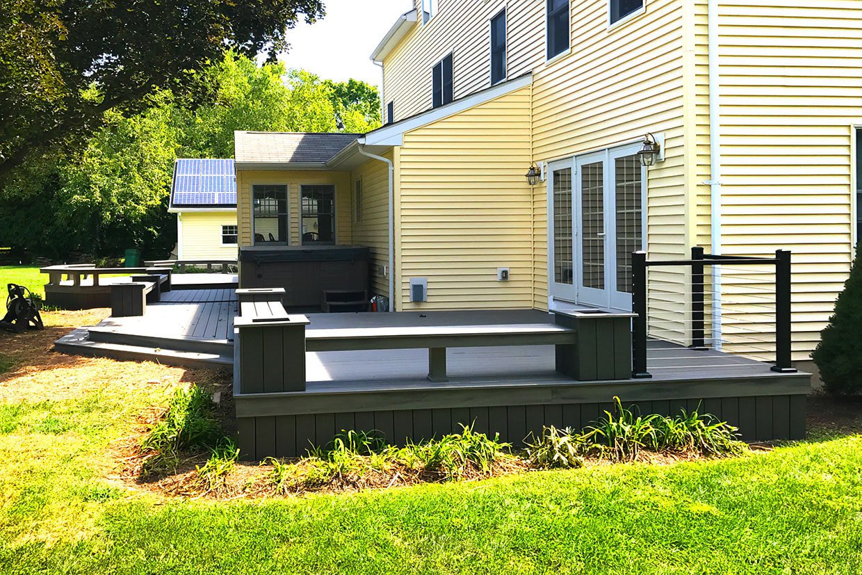A modern grey ground level deck with built-in bench seating, black cable railings, and a hot tub area.