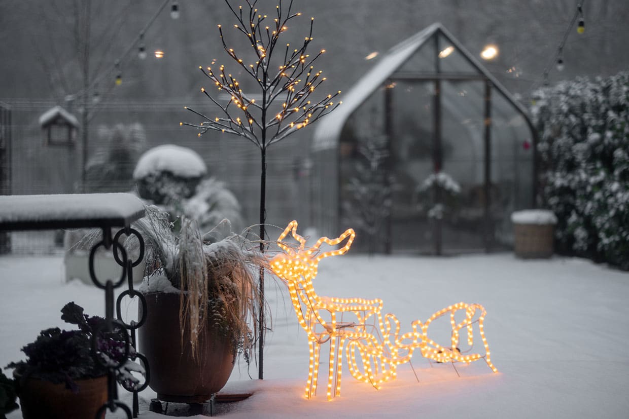 Glowing wire reindeer and lighted bare tree on a snow-covered patio, showcasing festive deck decorating ideas for Christmas.