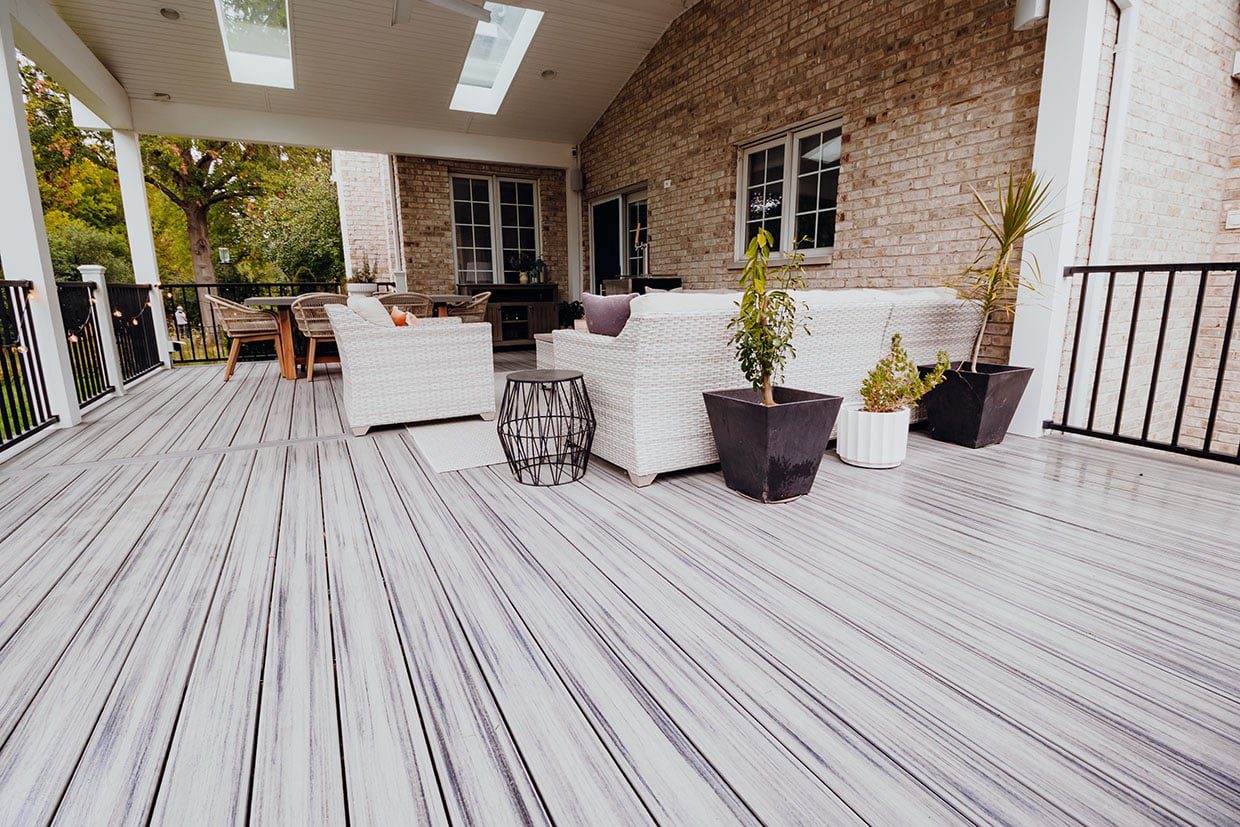 Close-up of gray composite decking colors with variegated streaking on a covered porch featuring white wicker furniture and a brick house wall.