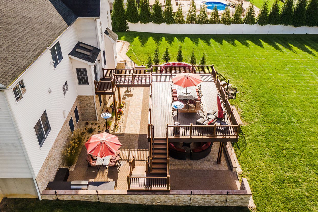 Aerial view of a large multi-level outdoor space showcasing expansive under deck landscaping ideas, featuring a composite upper deck and stone pavers creating a shaded patio zone.