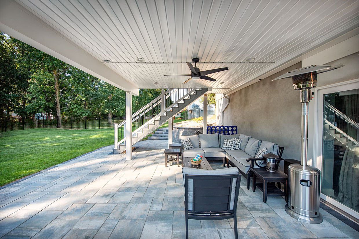 A modern under deck patio featuring grey stone pavers, a finished white ceiling with a fan, and a sectional sofa for outdoor living.