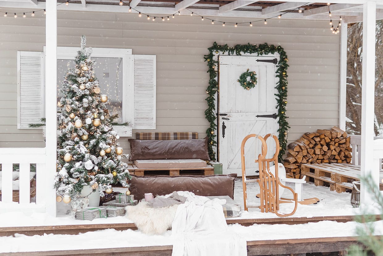 Covered deck featuring rustic Christmas deck decor, including a flocked Christmas tree, pallet seating with warm furs, a wooden sled, and garland framing a barn door under string lights.
