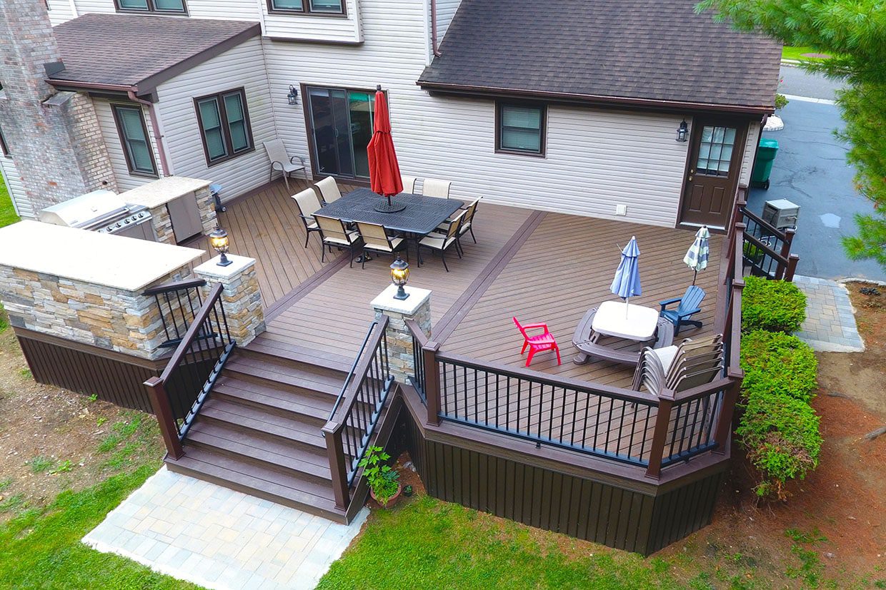 High-angle view of a multi-level deck featuring light brown composite decking colors with a dark brown picture frame border, an outdoor kitchen, and stone columns.