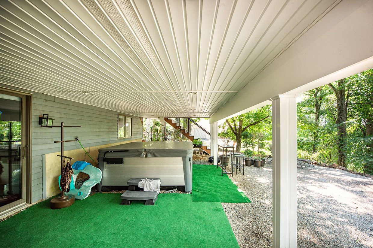 A dry under deck patio featuring a hot tub, artificial turf flooring, and a white ceiling drainage system, transitioning to a gravel landscape.