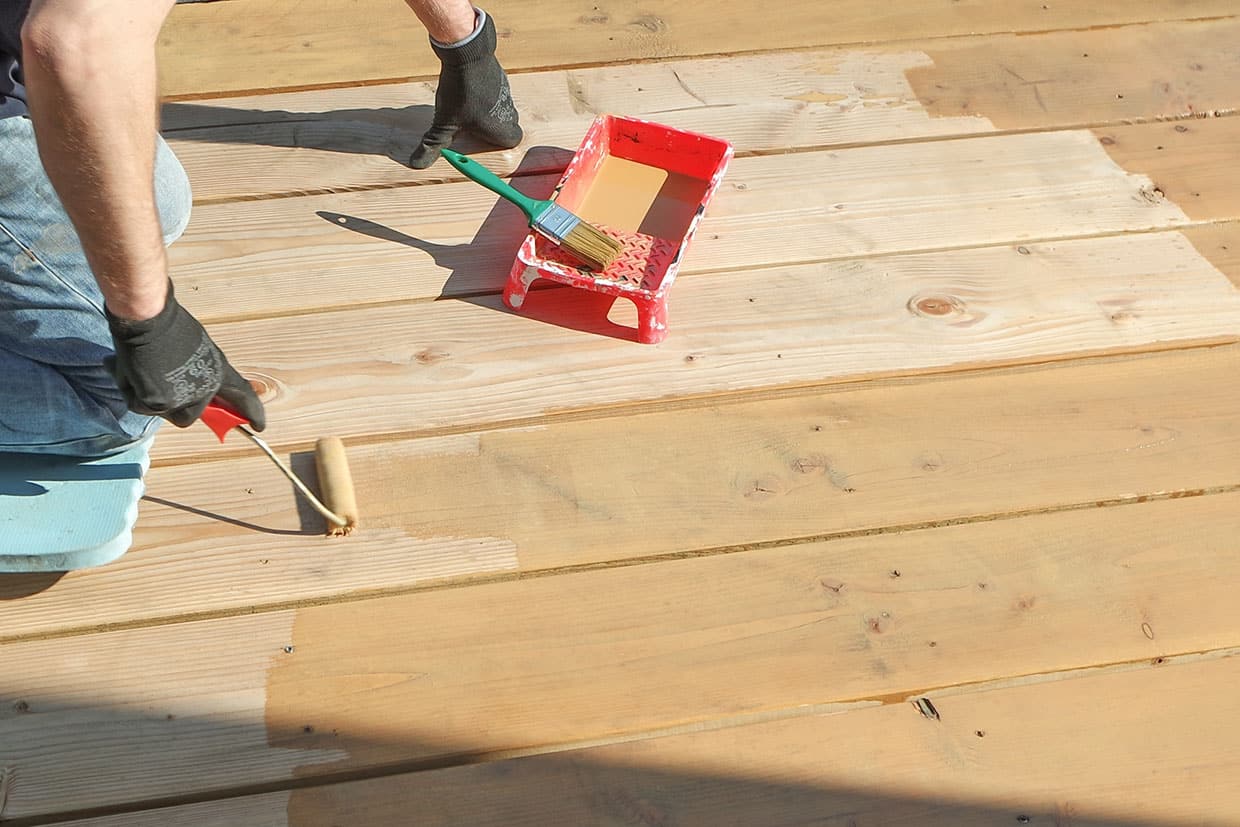 Worker applying a semi-transparent stain to raw wood deck boards using a small paint roller.