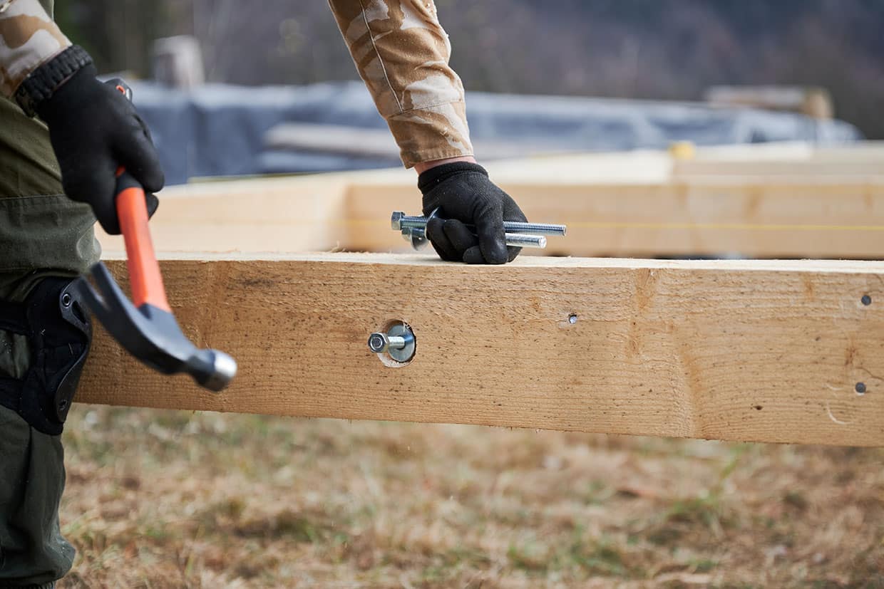 Close-up of a builder holding a hammer and galvanized carriage bolts against a wooden beam, illustrating hardware selection for a deck framing expert guide.
