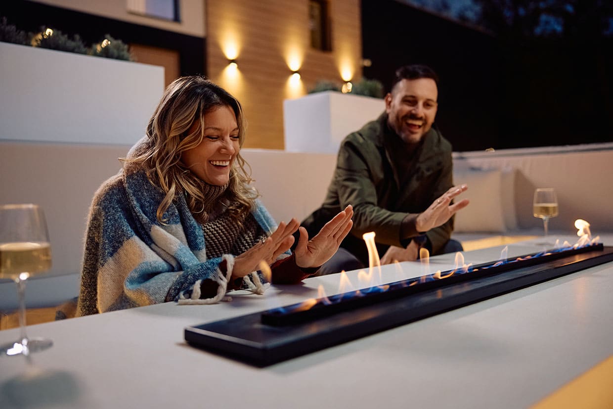 A happy couple warming their hands over a modern fire pit table, enjoying their cozy deck furnish arrangement at night.