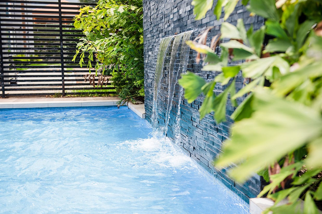 A modern stone wall with three waterfalls cascading into a blue swimming pool next to lush green plants.