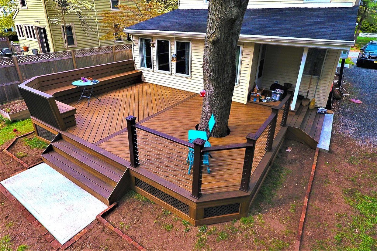 A custom pet deck with rich brown composite flooring, cable railing, built-in benches, and a large tree at its center.