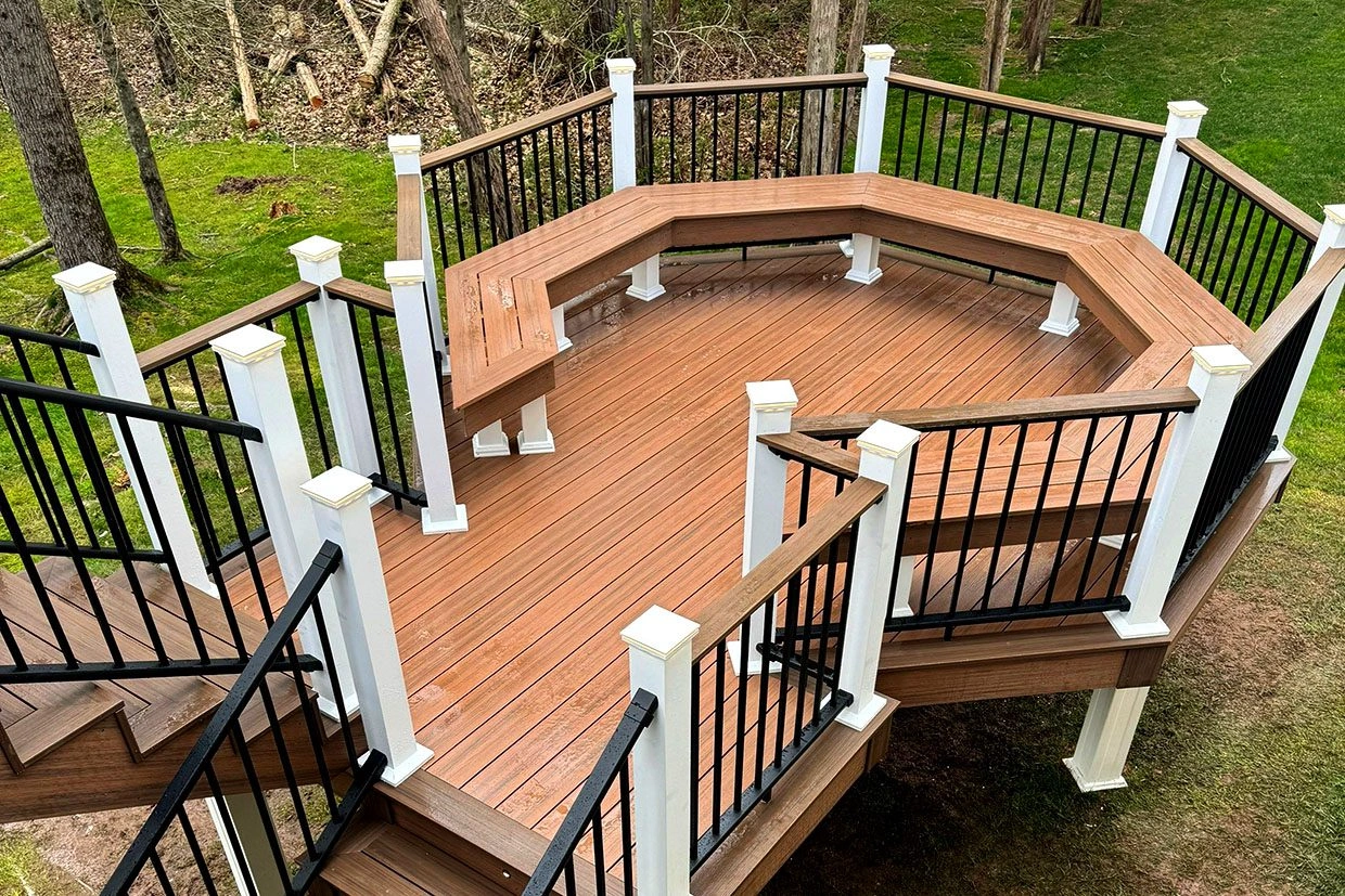 Overhead view of a new octagonal deck with integrated wood-tone bench, white posts, and black railings surrounded by trees and lawn where to find reliable reviews about deck companies.