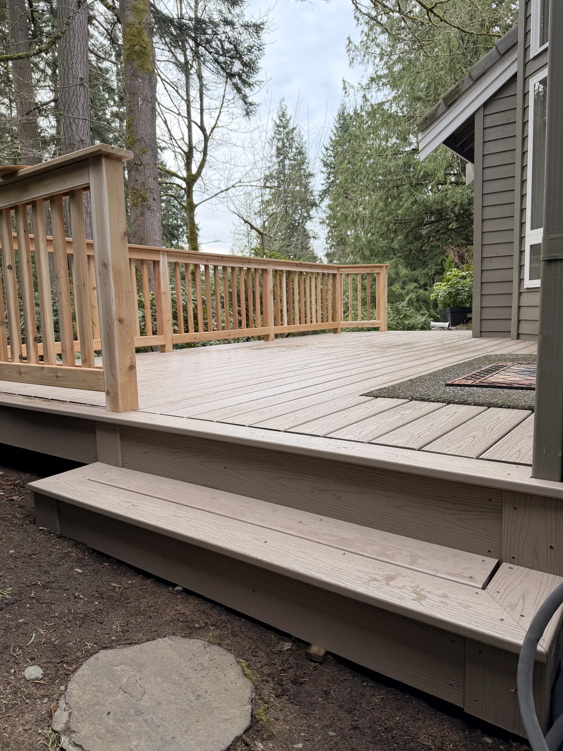 Complete Composite Deck Installation in Mill Creek WA with Cedar Railings A finished Composite Deck Installation in Mill Creek WA with light gray planks and natural wood railings next to a house and forest.