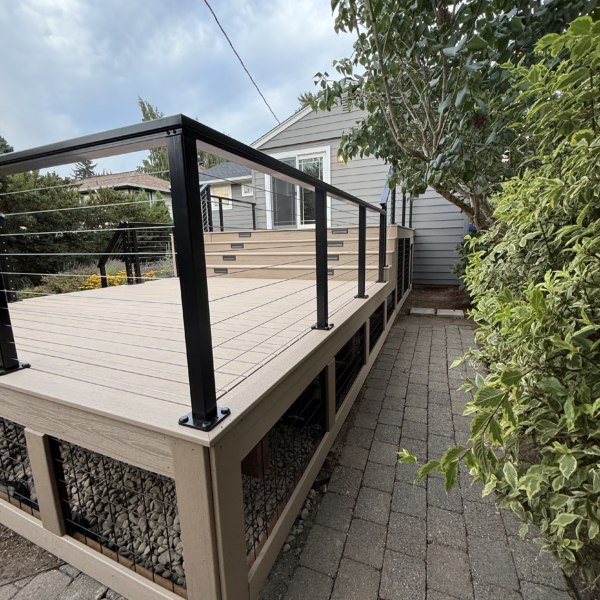 Second-story Trex deck replacement Side Profile Of A Composite Deck Showing Black Cable Railing Posts And Wire Mesh Under-Deck Fencing Along A Paver Path.
