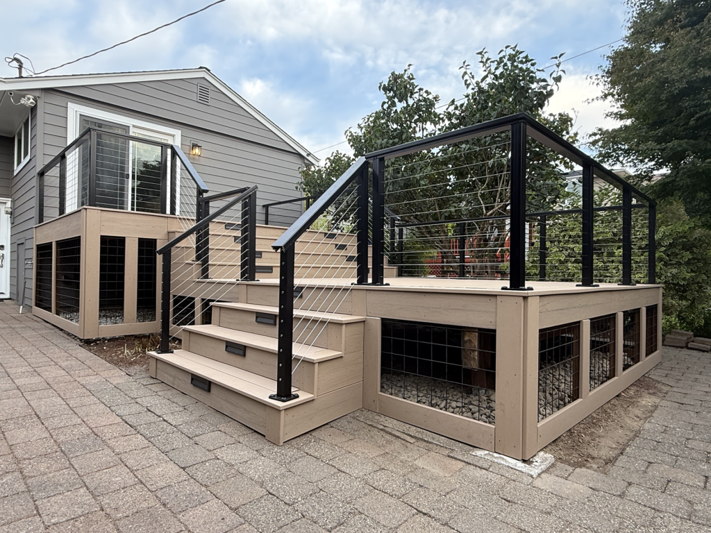 New Decks Multi-Level Beige Composite Deck With Black Cable Railings And Under-Deck Wire Mesh Skirting Next To A Paver Patio.
